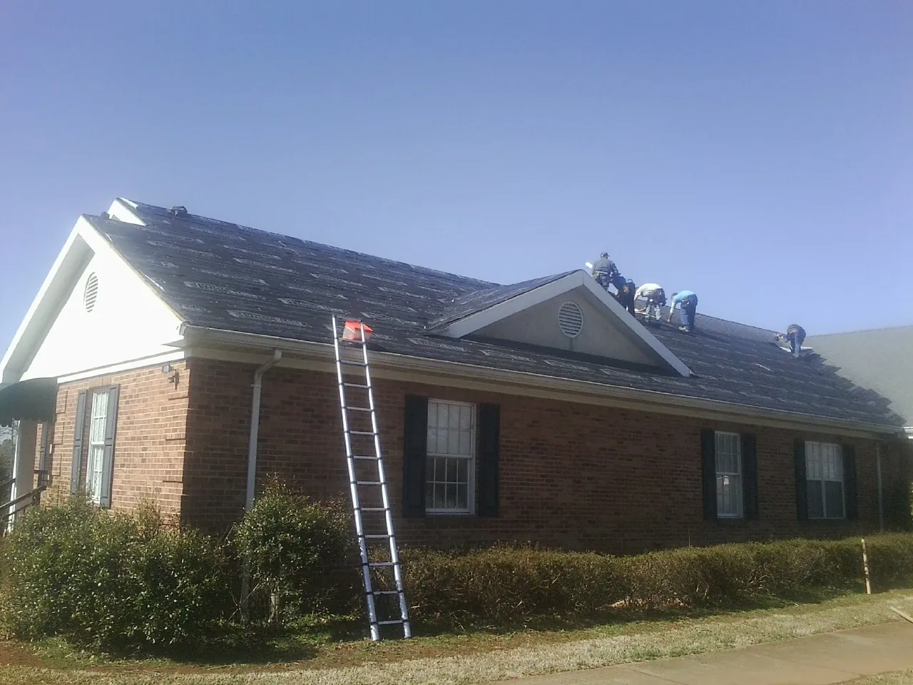 Crew tearing shingles off a brick ranch roof with a ladder set — Emergency Roof Repair work by our crew in Melville