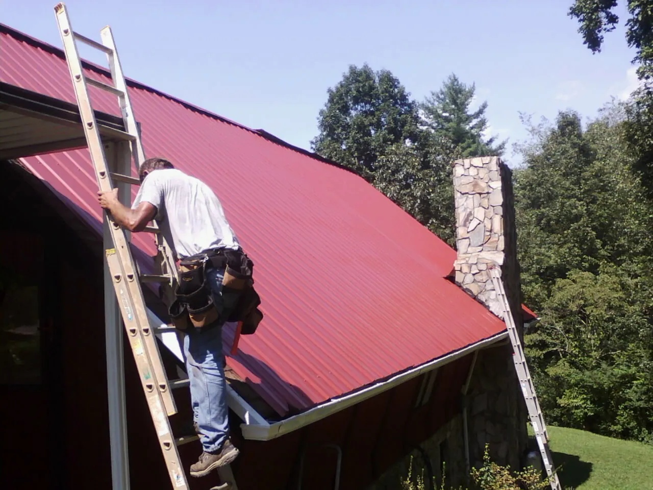 Roofer on a ladder beside a red metal roof and stone chimney — Soffit & Fascia Repair by Roofing Brothers in Melville